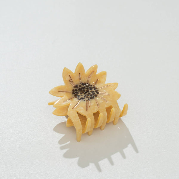 A small sunflower-shaped hair claw clip in a warm pale yellow tone, with a dark brown patterned center and thin gold metallic rays, photographed on a clean white background.