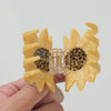 A hand holding a yellow sunflower-shaped hair claw clip with a clear plastic hinge and brown polka dot pattern on the inner petals, against a plain light gray background.