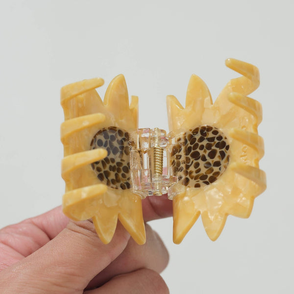 A hand holding a yellow sunflower-shaped hair claw clip with a clear plastic hinge and brown polka dot pattern on the inner petals, against a plain light gray background.