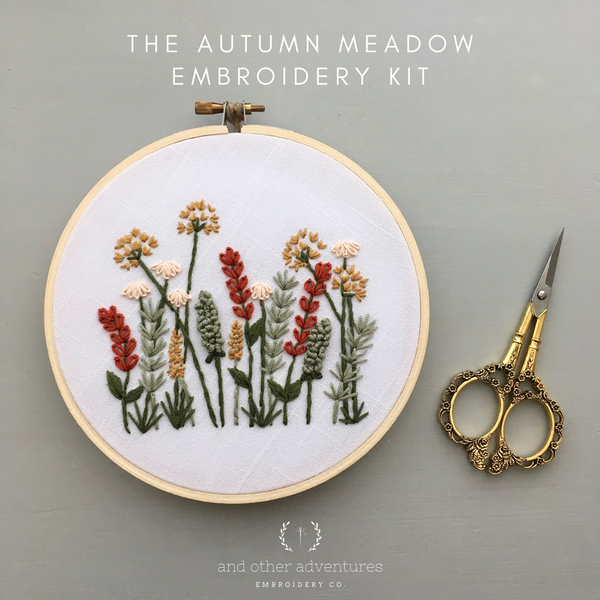 An embroidery kit display featuring a wooden hoop with a white fabric and embroidered wildflowers in red, beige, green, and gray, alongside ornate gold scissors on a gray background. Text reads 'THE AUTUMN MEADOW EMBROIDERY KIT' and 'and other adventures EMBROIDERY CO.