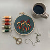 A styled flat-lay of embroidery supplies on a light gray surface, featuring a round wooden hoop with a floral needlepoint or embroidery design in red, orange, and cream threads on a dark green background. Surrounding the hoop are five spools of embroidery floss in peach, rust, green, and olive tones, a pair of ornate metal embroidery scissors, loose green thread, and a gray ceramic mug filled with a dark beverage.