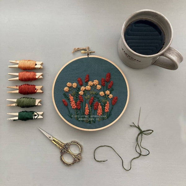 A styled flat-lay of embroidery supplies on a light gray surface, featuring a round wooden hoop with a floral needlepoint or embroidery design in red, orange, and cream threads on a dark green background. Surrounding the hoop are five spools of embroidery floss in peach, rust, green, and olive tones, a pair of ornate metal embroidery scissors, loose green thread, and a gray ceramic mug filled with a dark beverage.
