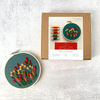 A flat-lay product shot of a floral embroidery kit, showing a completed round embroidery hoop with red and cream flowers on a dark green background next to its brown cardboard packaging box.