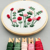 A round embroidery hoop holding white fabric with a floral stitched design in red, pink, and green thread, shown on a light gray surface. Five spools of embroidery thread in green, white, pink, and red are aligned along the bottom, with the text 'AND OTHER ADVENTURES EMBROIDERY CO' visible.