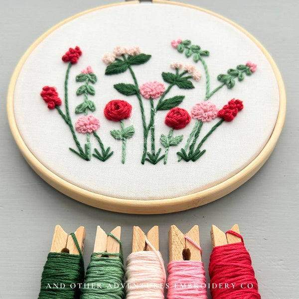 A round embroidery hoop holding white fabric with a floral stitched design in red, pink, and green thread, shown on a light gray surface. Five spools of embroidery thread in green, white, pink, and red are aligned along the bottom, with the text 'AND OTHER ADVENTURES EMBROIDERY CO' visible.