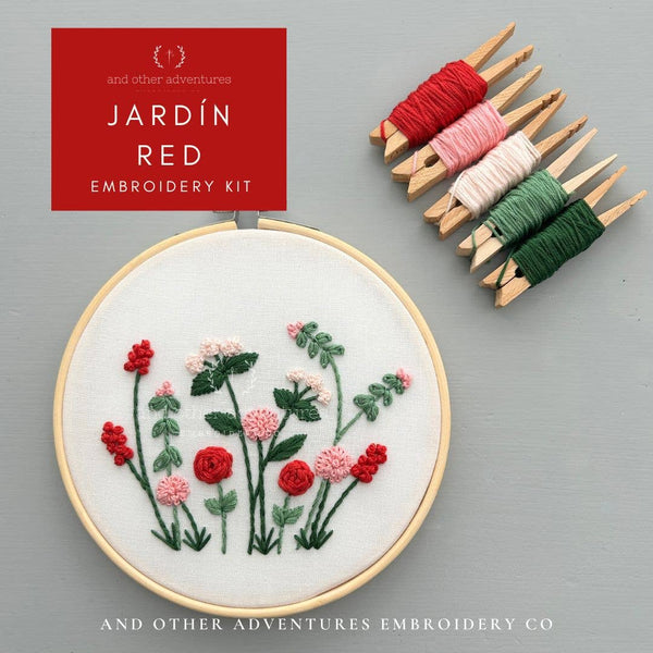 Embroidered floral kit shown in a wooden hoop with red and pink flowers and green leaves on white fabric, alongside five thread spools in red, pink, white, and green. A red label reads 'JARDIN RED EMBROIDERY KIT' and the brand 'And Other Adventures Embroidery Co' appears at the bottom.