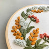 Close-up of a circular embroidery hoop holding cream fabric with embroidered flowers and leaves in orange, coral pink, pale blue-gray, and olive green.