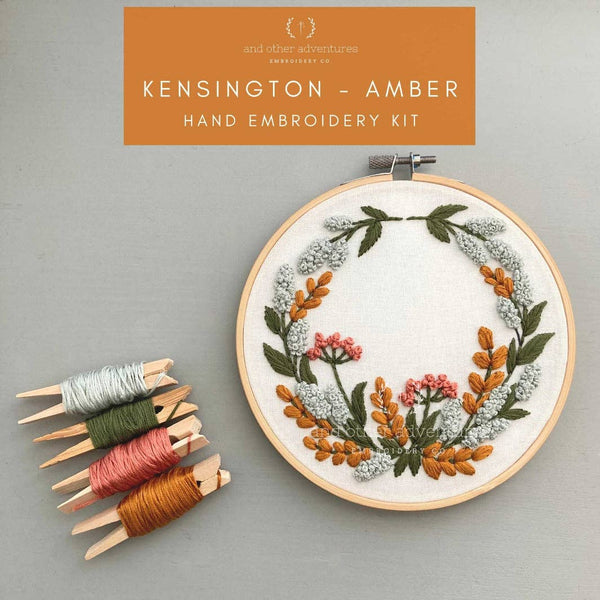 A hand embroidery kit shown with a wooden hoop containing a floral wreath design, several thread spools on wooden clips, and an orange label reading 'KENSINGTON - AMBER HAND EMBROIDERY KIT' on a light gray background.