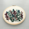 A round embroidery hoop holding cream fabric with a floral needlepoint design in muted green, dusty pink, and burgundy tones, photographed on a light gray background. Text at the bottom reads 'AND OTHER ADVENTURES EMBROIDERY CO'.