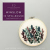 A hand embroidery kit preview showing a wooden hoop with embroidered wildflowers in muted green, blue, burgundy, and pink on cream fabric, alongside a purple brand label reading 'Winslow in Spellbound Hand Embroidery Kit' and 'And Other Adventures Embroidery Co'.