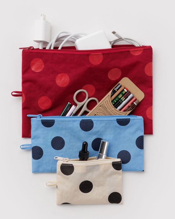 Three fabric zip pouches with polka dots are arranged on a light gray background. The top pouch is red with red polka dots and contains white charging accessories. The others include various pens, pencils, scissors, and other small items. 