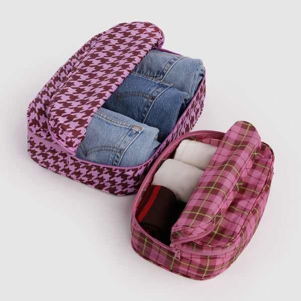 Pink houndstooth and plaid-patterned travel organizers with denim jeans and white socks on a light gray background