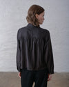 A person facing away toward a wall showing the back of a dark brown and black striped blouse worn over black pants. 