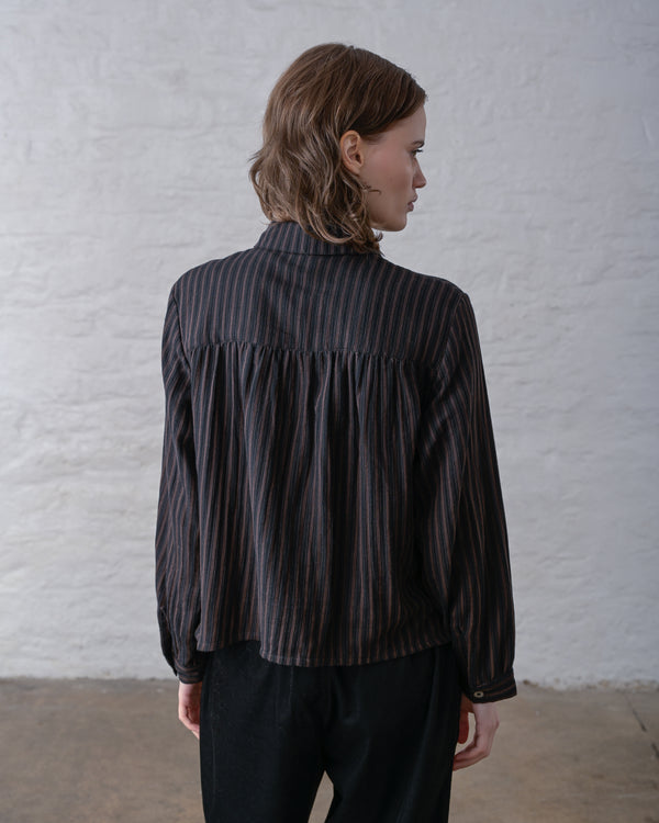 A person facing away toward a wall showing the back of a dark brown and black striped blouse worn over black pants. 