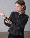 A person doing up the sleeve cuff button on a dark brown and black striped button up blouse, styled with black pants, as they stand in front of a white wall.