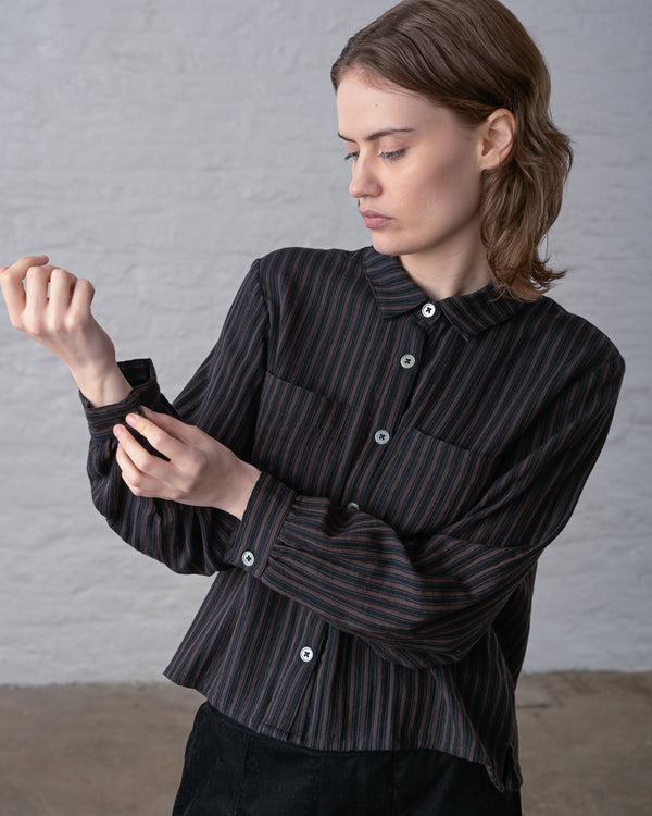 A person doing up the sleeve cuff button on a dark brown and black striped button up blouse, styled with black pants, as they stand in front of a white wall.