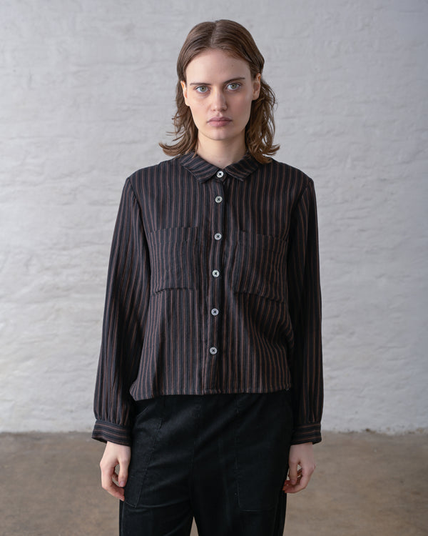 Person wearing a dark brown and black striped button up shirt with black pants against a plain background.