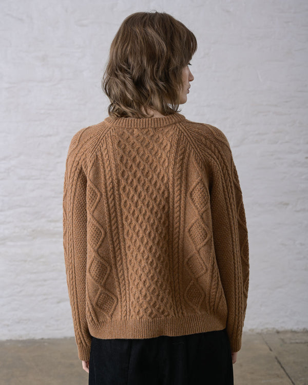 A person facing a white wall wearing an almond brown Aran cable knit cardigan over black pants.