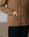 A close-up view of a person with their hand in the pocket of an almond-brown cable knit cardigan, worn over black pants.