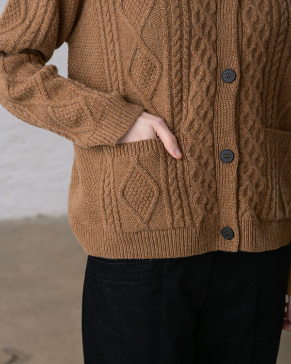 A close-up view of a person with their hand in the pocket of an almond-brown cable knit cardigan, worn over black pants.