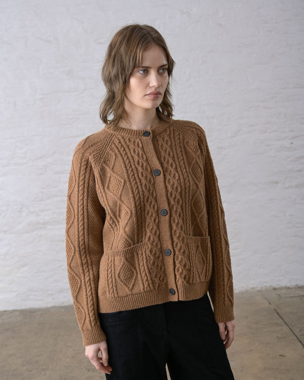 A person standing against a white wall wearing an almond brown Aran cable knit cardigan with pockets over black pants.