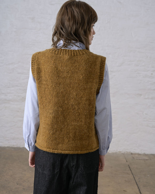 A person facing away toward a white wall, wearing a bronze handknit sweater vest styled over a light blouse and black pants.
