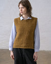 A person wearing a bronze handknit sweater vest styled over a light blouse and black pants, standing against a white wall.