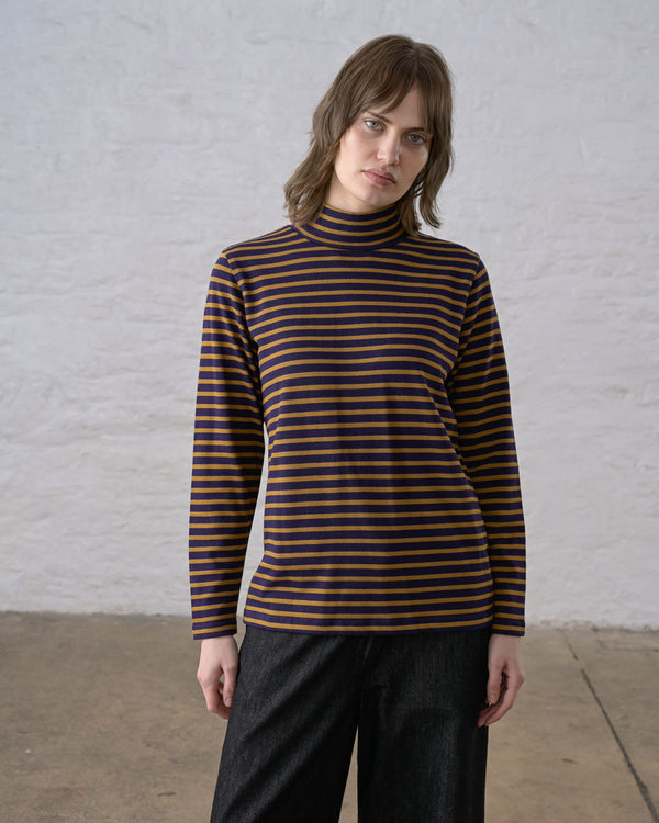 A person standing against a white wall wearing a long sleeve dark navy and bronze striped knit top with a mockneck collar, styled over black pants.