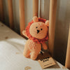 A crocheted lion rattle toy with an orange body and rust-orange mane sits on a white quilted mattress next to wooden crib rails. A paper tag attached to the toy reads 'BITSY ZOO'