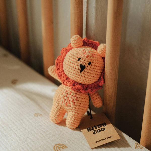 A crocheted lion rattle toy with an orange body and rust-orange mane sits on a white quilted mattress next to wooden crib rails. A paper tag attached to the toy reads 'BITSY ZOO'