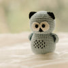 A small crocheted owl rattle toy in light gray with black ears, black eyes, a black nose, and cream-colored eyes, standing on a soft white fabric surface.