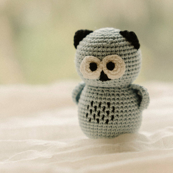 A small crocheted owl rattle toy in light gray with black ears, black eyes, a black nose, and cream-colored eyes, standing on a soft white fabric surface.