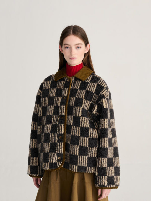 A person wearing a beige and grey striped checkerboard fleece jacket with brown corduroy trim and collar over a red turtleneck and brown skirt against a white background.