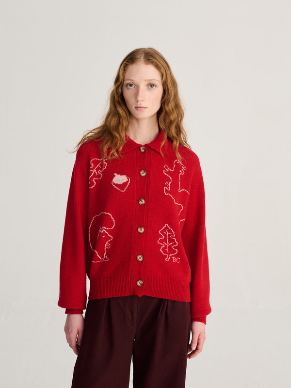 A model wearing a red sweater with a white forest scene and dark red pants against a white wall.