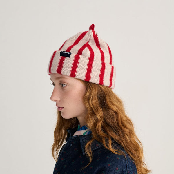A person wearing a red and white striped wool beanie hat with a red top and a small black 'Bobo Choses' label along with a dark denim floral print shirt against a white background.