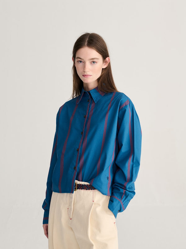 A person wearing a bold blue and red triple stripe poplin collared shirt and cream colored pants against a white background.