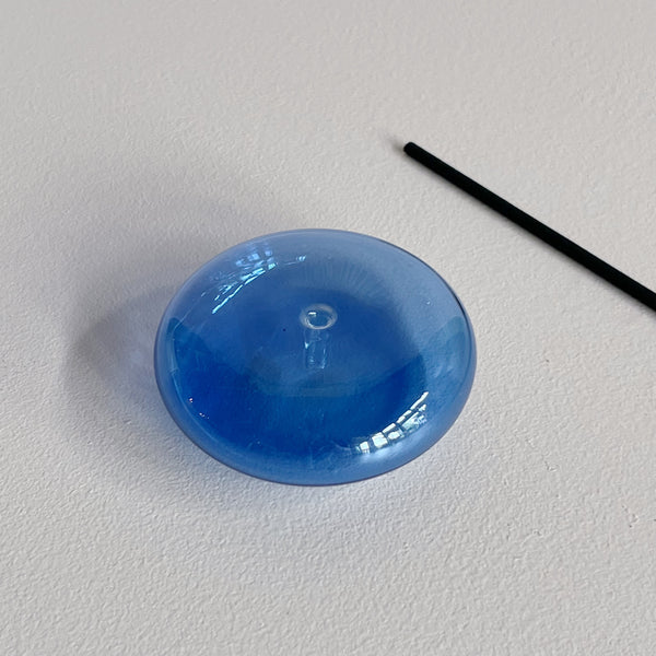 A translucent blue bubble-shaped glass incense holder with a stick of incense laying next to it against a neutral background.