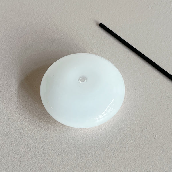 A white milk glass bubble-shaped incense holder with a stick of incense lying. next to it against a neutral background.