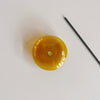 A close top-down view of a translucent amber bubble glass incense holder with a stick of incense lying next to it on a neutral background.