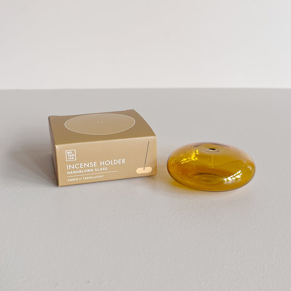 A translucent amber glass bubble shaped incense holder sitting next to its packaging on a neutral backdrop.