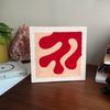 A square abstract wall art piece with a white frame sits on a wooden desk. The punch needle artwork features a bold red abstract shape on a peach-pink textured background.