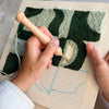 A close-up of a person's hands using a wooden-handled punch needle to weave a textured art piece in dark green and light sage green on a beige canvas.