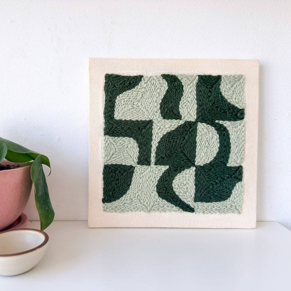 A square abstract needlepoint canvas with dark green and light sage green organic shapes on a cream border, displayed on a white surface next to a pink potted plant and a small ceramic bowl.