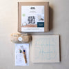 A flat-lay view of a 'Both Hands' Punch Needle Kit on a textured beige background, showing the brown cardboard box, a wrapped bundle, a printed instruction sheet, and a canvas with blue pattern.