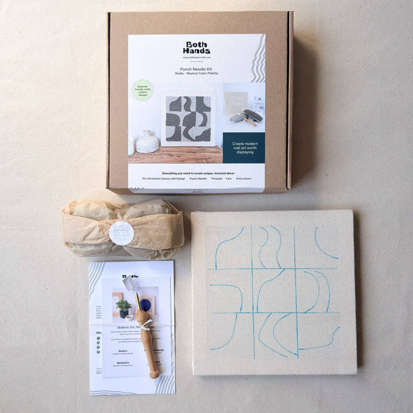 A flat-lay view of a 'Both Hands' Punch Needle Kit on a textured beige background, showing the brown cardboard box, a wrapped bundle, a printed instruction sheet, and a canvas with blue pattern.