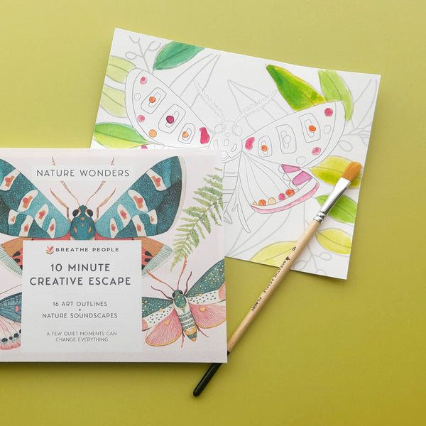 A half-painted outlined page of a butterfly with a wooden paintbrush and the 'Nature Wonders' edition of the '10 Minute Creative Escape' art book by Breathe People against a bright green background.