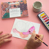 A top-down view of a watercolor painting setup on a pink background. A hand holds a paintbrush over a white paper sketch of a flower, with a watercolor palette, a small cup of water, and a book labeled 'Vintage Blooms 10 Minute Creative Escape' nearby.