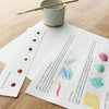 Instructional materials on watercolor techniques with a cup and brush on a wooden surface.