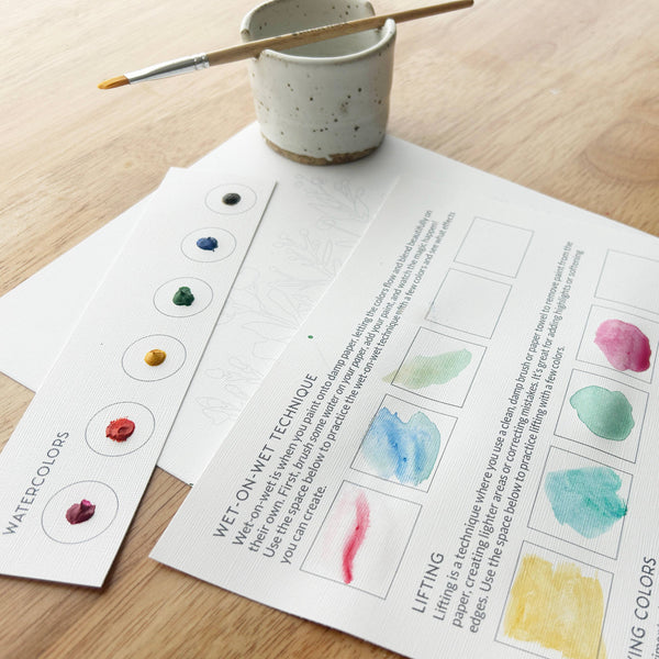 Instructional materials on watercolor techniques with a cup and brush on a wooden surface.