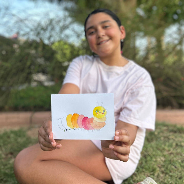 Kid holding a watercolor painted postcard of a caterpillar while sitting outside.